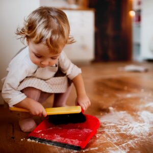 A small toddler girl with brush and dustpan sweeping floor in the kitchen at home.